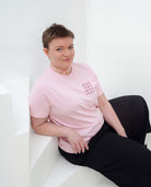 Model wearing light pink Biweekly T-shirt with clit graphic print on right chest area, front view, sitting on stairs against white background
