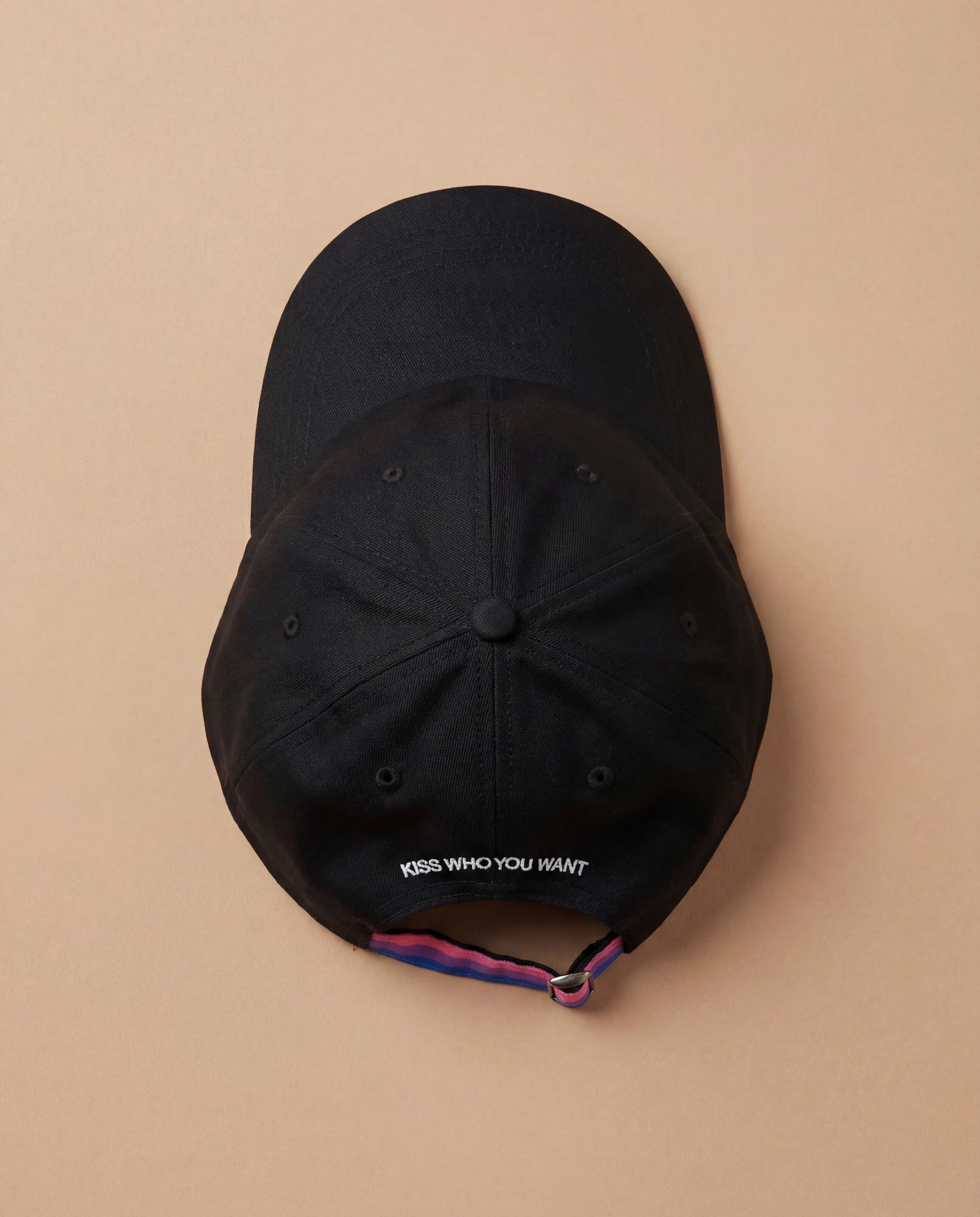 Black Biweekly cap with a strap in the colors of the bi pride flag and the text ‘Kiss Who You Want’ on the back, laying flat on beige background