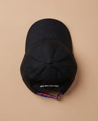 Black Biweekly cap with a strap in the colors of the bi pride flag and the text ‘Kiss Who You Want’ on the back, laying flat on beige background