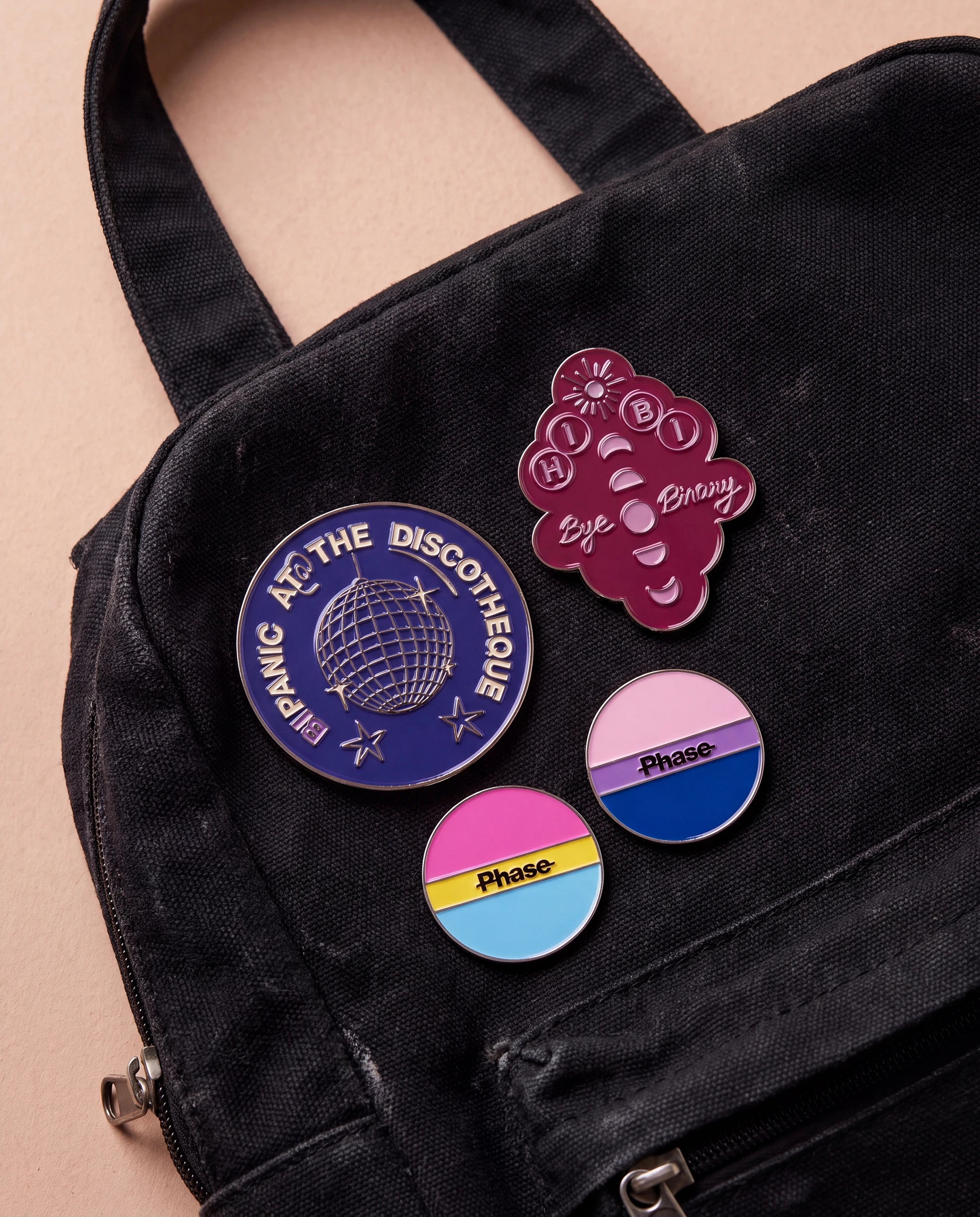 Burgundy 'Hi Bi, Bye Binary' pin, bi coloured 'Phase' pin, pan coloured 'Phase' pin and blue 'Bipanic at the discotheque' pin are displayed. All four pins are placed on a black bag.