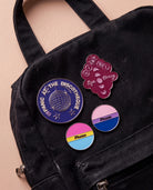 Burgundy 'Hi Bi, Bye Binary' pin, bi coloured 'Phase' pin, pan coloured 'Phase' pin and blue 'Bipanic at the discotheque' pin are displayed. All four pins are placed on a black bag.