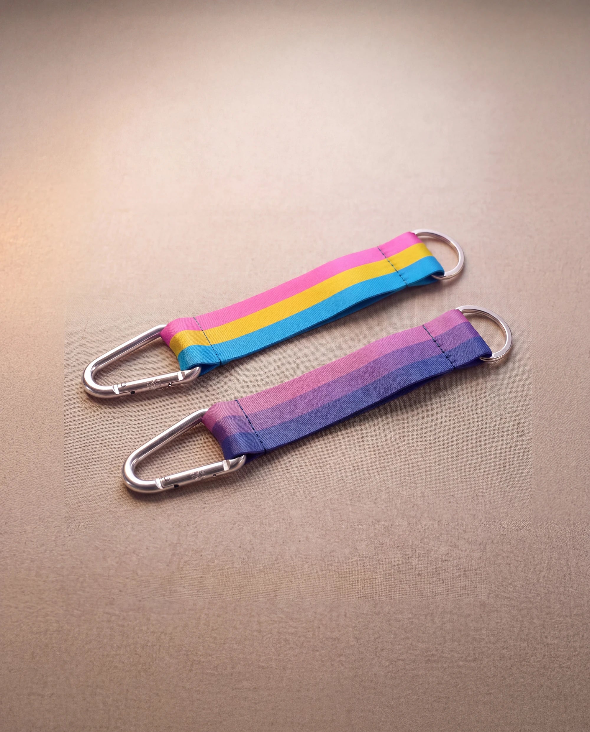 Two Keychain in bi pride and pan pride flag colors with silver carabiner and blue yarn laying next to eachother against beige background