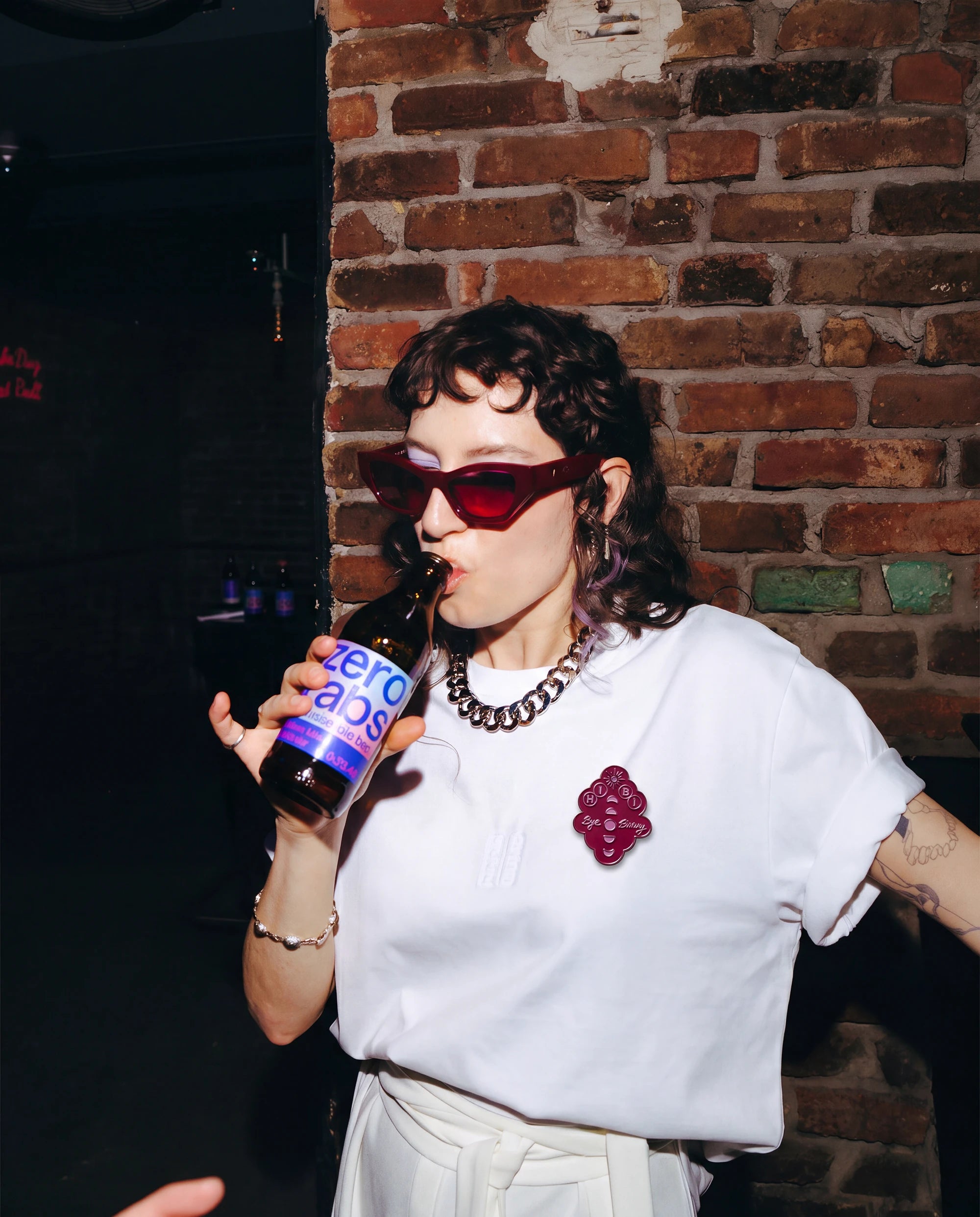 Model wearing burgundy Biweekly pin with 'Hi Bi - Bye Binary' text and moon phase illustration on a white Biweekly Shirt. 
