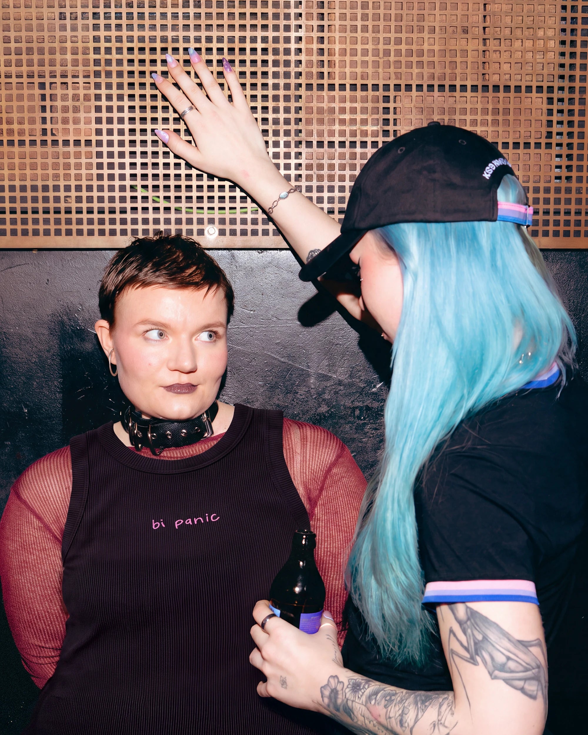 Model wearing a black Biweekly tank top with pink ‘bi panic’ text, front view looking at another model which is seen from the back in a club setting