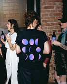 Model wearing an oversized black Biweekly T-shirt with a moon phase print that has gradient bi pride colours, back view in a club setting