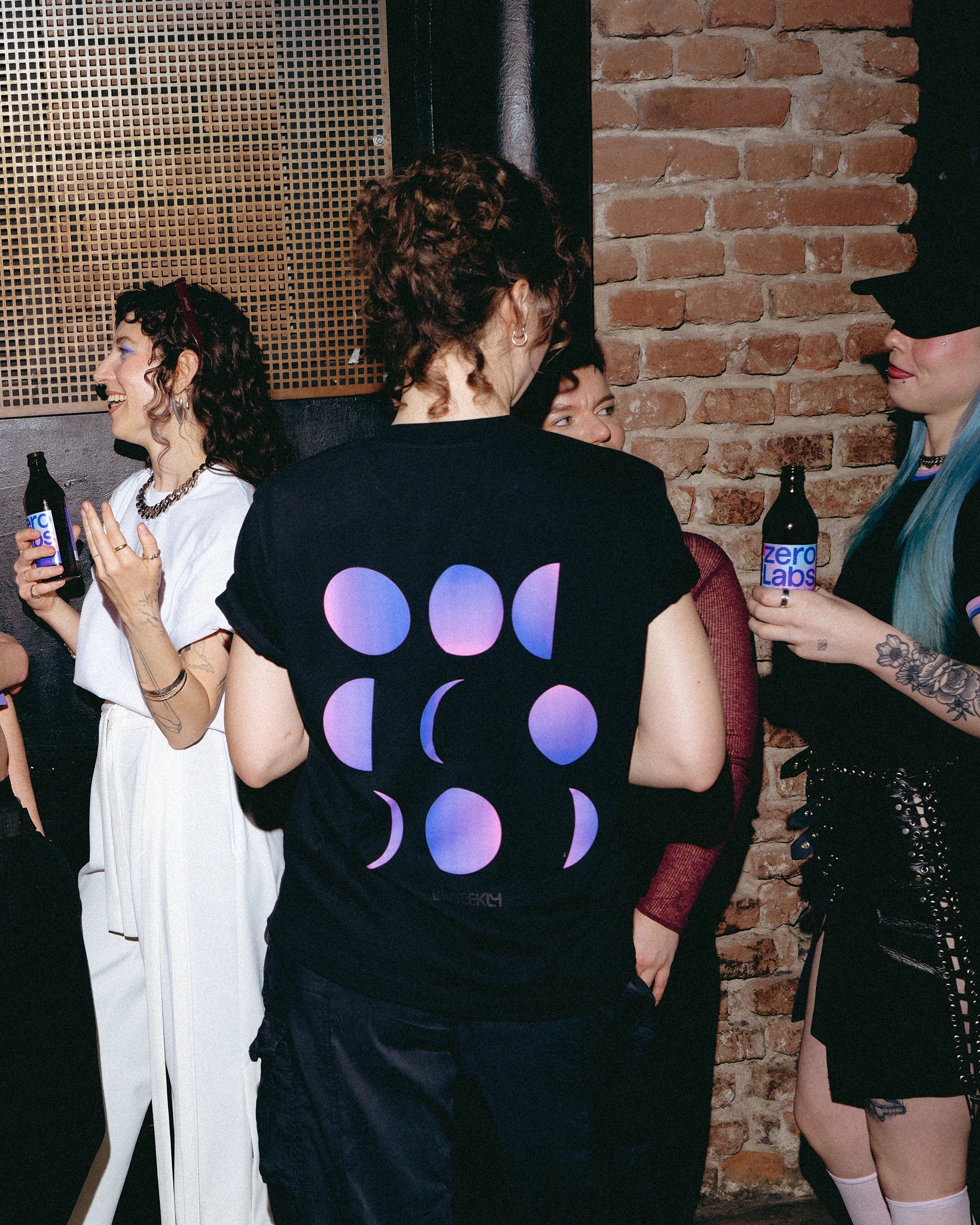 Model wearing an oversized black Biweekly T-shirt with a moon phase print that has gradient bi pride colours, back view in a club setting