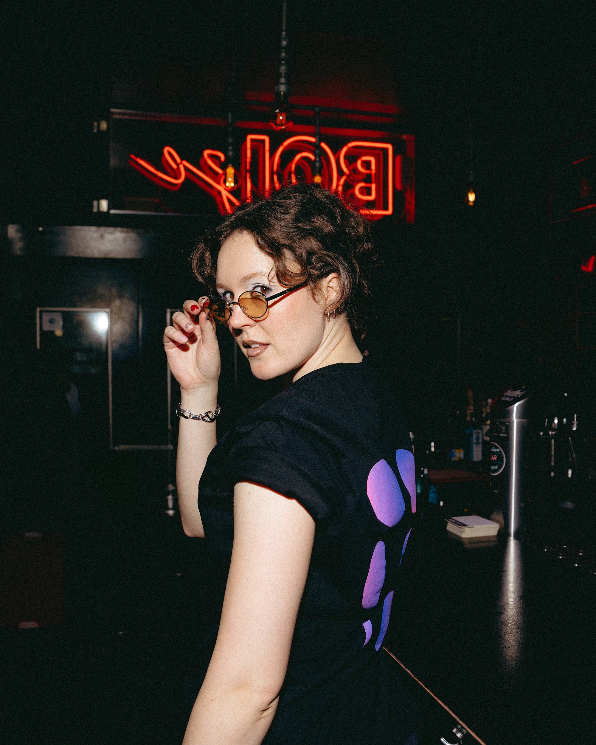Model wearing an oversized black Biweekly T-shirt with a moon phase print that has gradient bi pride colours, back view in a club setting