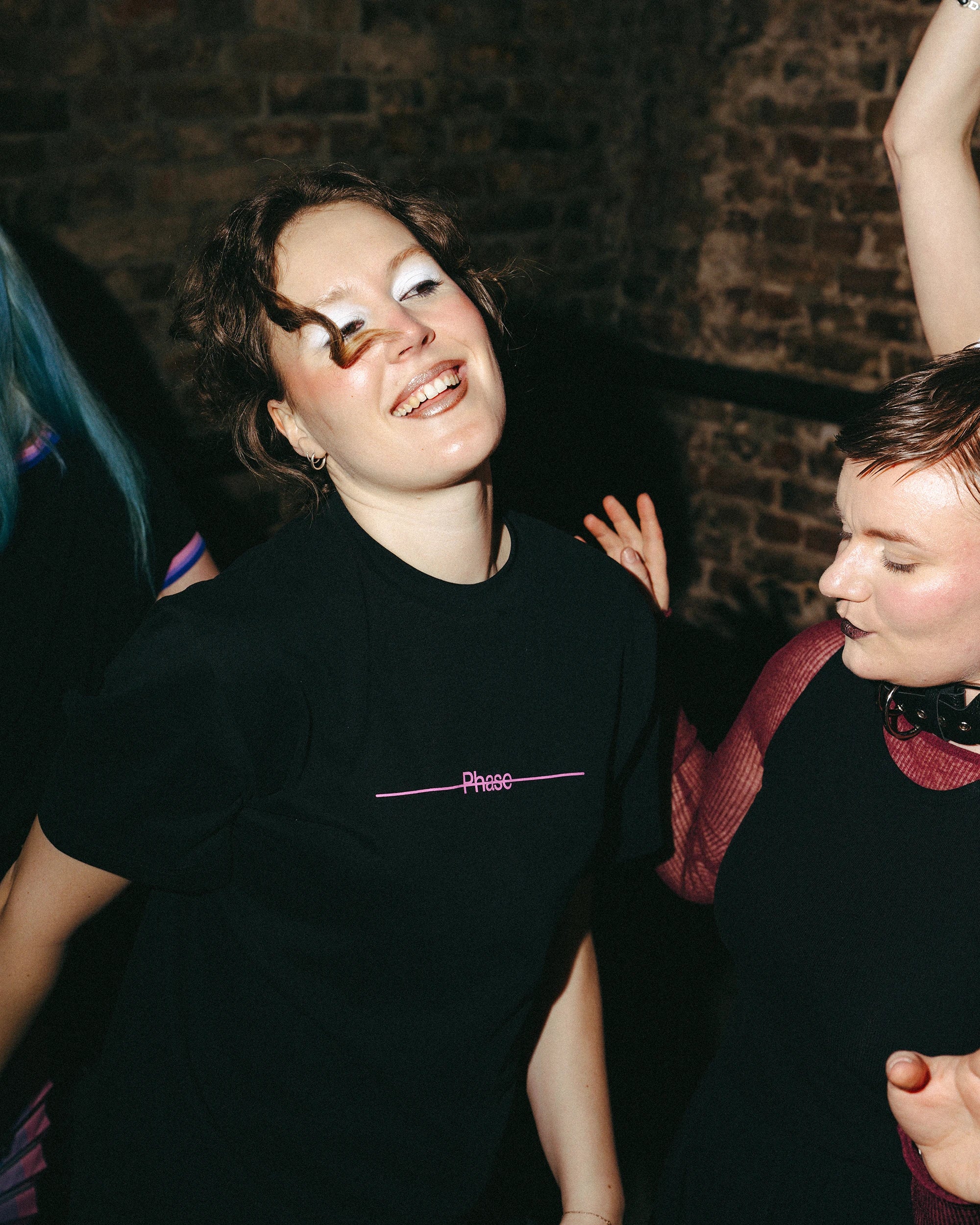Model wears an oversized black Biweekly T-shirt with pink text 'Phase' crossed out by a long line, front view dancing in a club scenario 