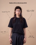 Model wears an oversized black Biweekly T-shirt with white ‘BiBi’ logo, front view against a beige background. With text that gives information on the shirt. 