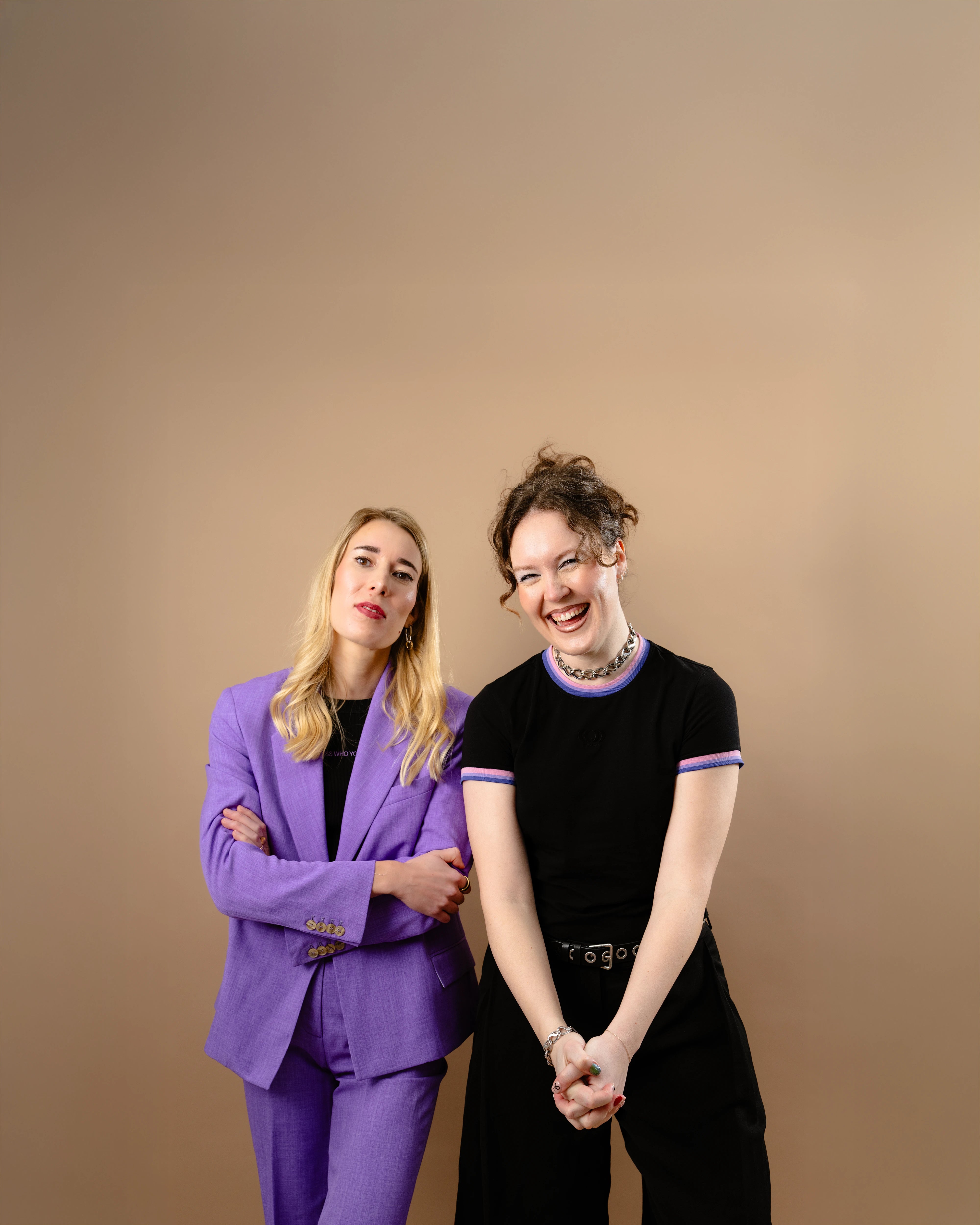 Two models standing next to each other wearing Biweekly Babytee and black croptop with lilac blazer on top, right model smiling and left model looking serious on beige bakground