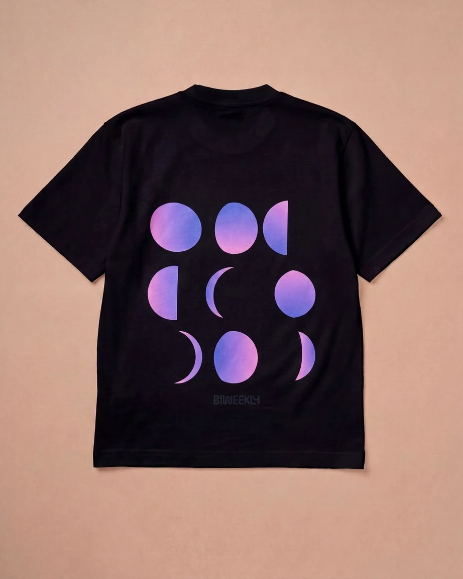 Oversized black Biweekly T-shirt with a moon phase print that has gradient bi pride colours, laying flat against beige background