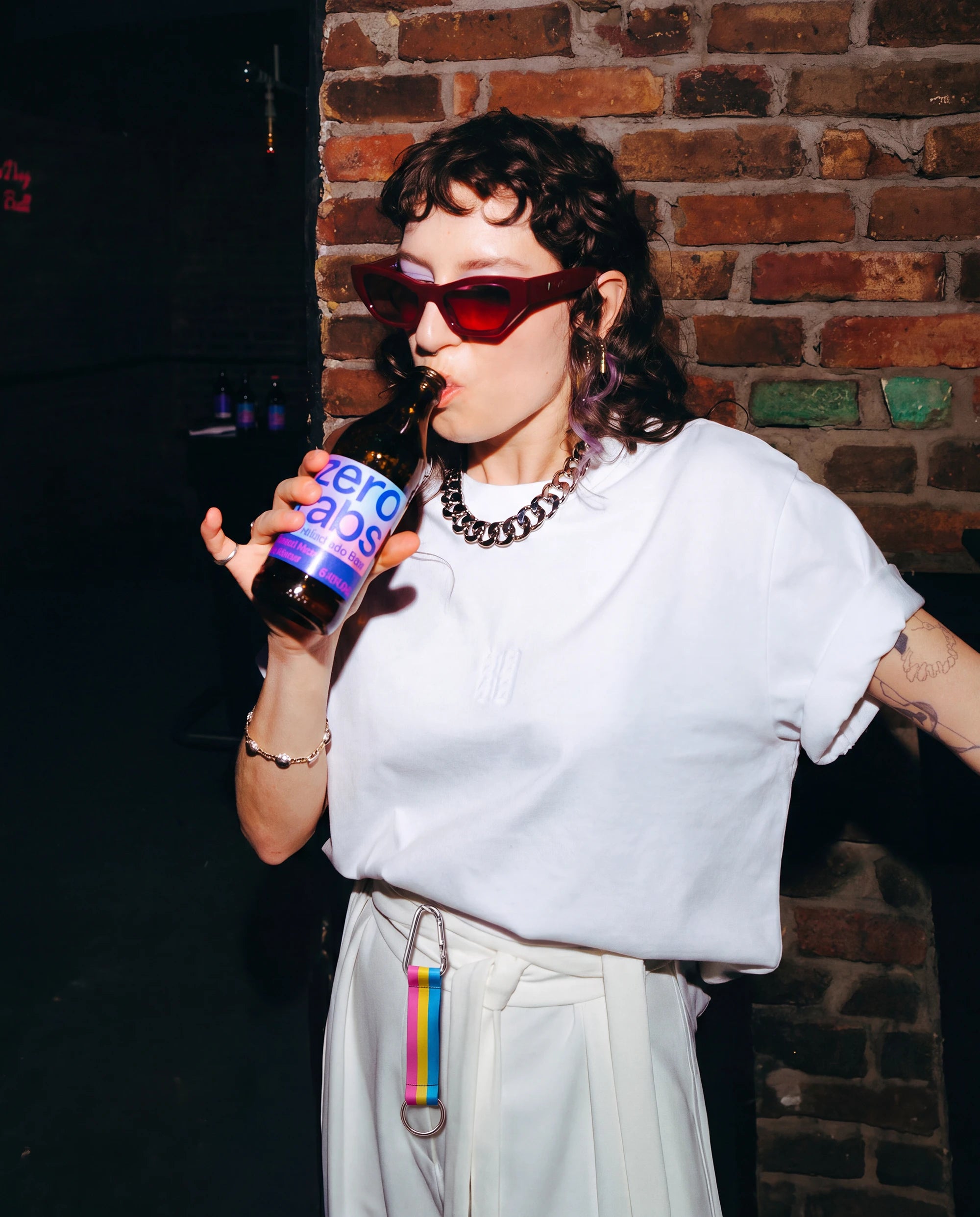 Model wears Biweekly keychain in pan pride flag colors with silver carabiner and blue yarn on skirt loops while drinking in a club scenario