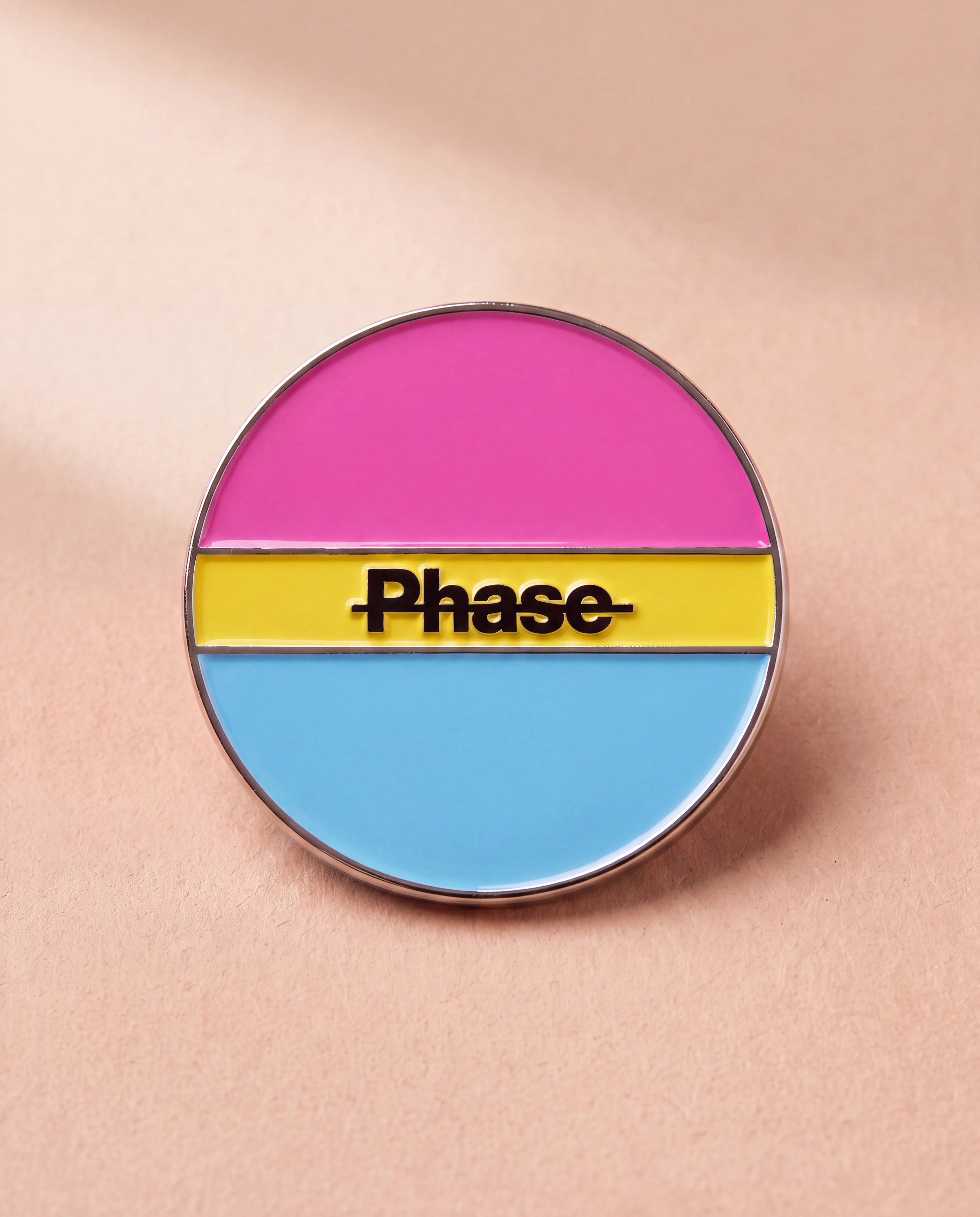 Biweekly Pin in pan colours with a crossed out 'Phase' text, flatlay against beige background