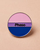 Biweekly Pin in bi colours with a crossed out 'Phase' text, flatlay against beige background