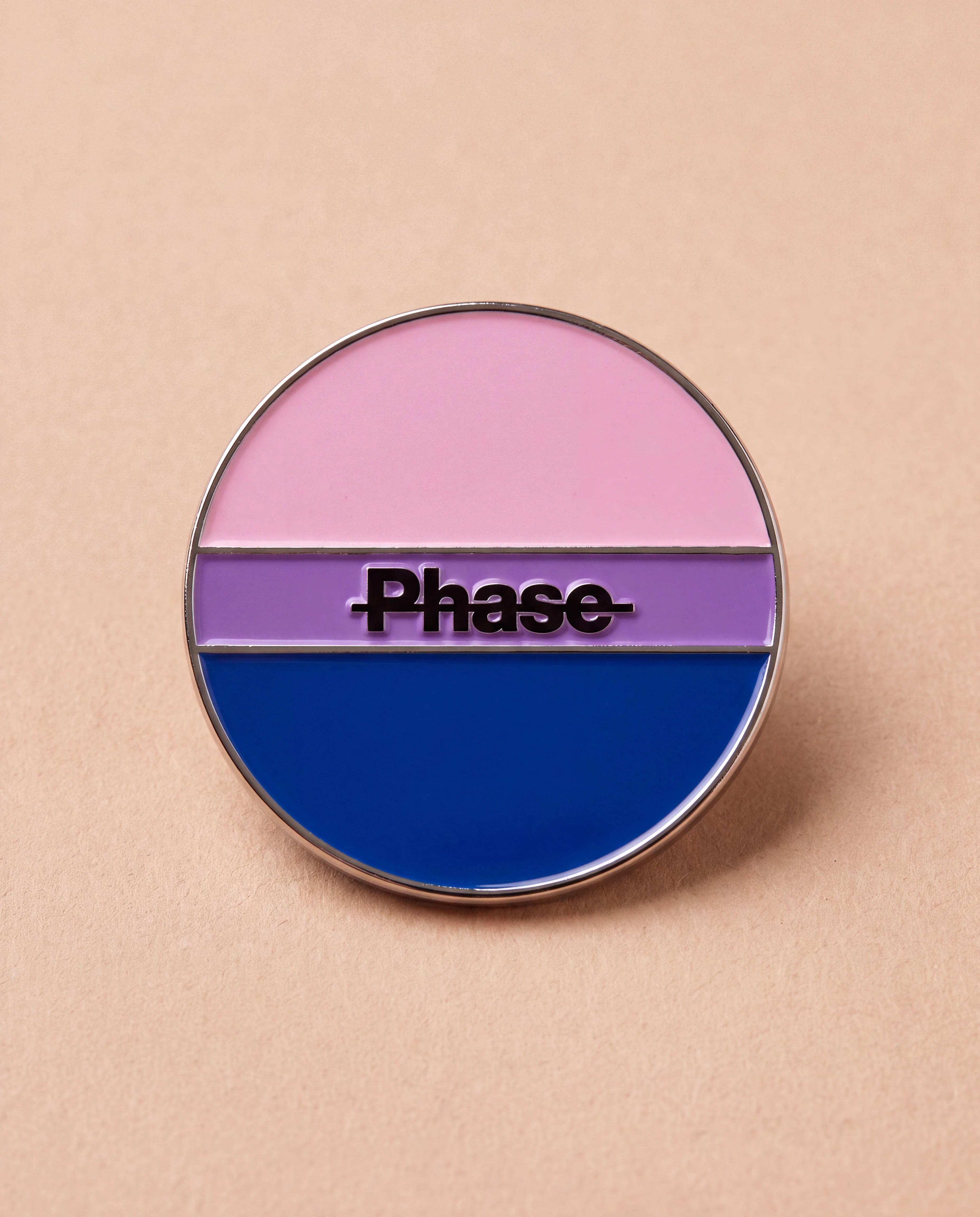 Biweekly Pin in bi colours with a crossed out 'Phase' text, flatlay against beige background
