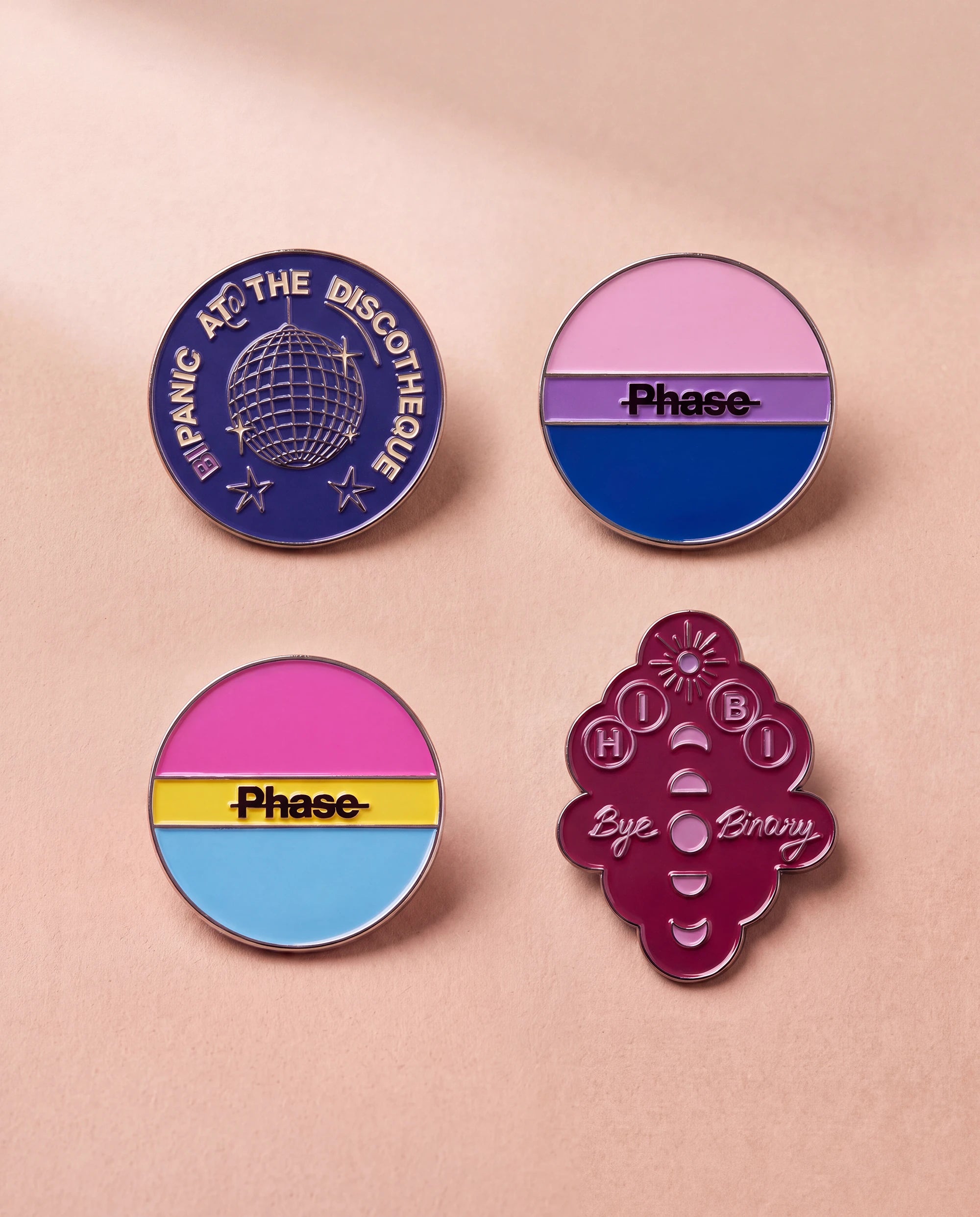 Burgundy 'Hi Bi, Bye Binary' pin, bi coloured 'Phase' pin, pan coloured 'Phase' pin and blue 'Bipanic at the discotheque' pin lay flat against beige background. 