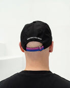 Model wearing a black Biweekly cap with bi pride flag strap and ‘Kiss Who You Want’ text on the back, back view against white background