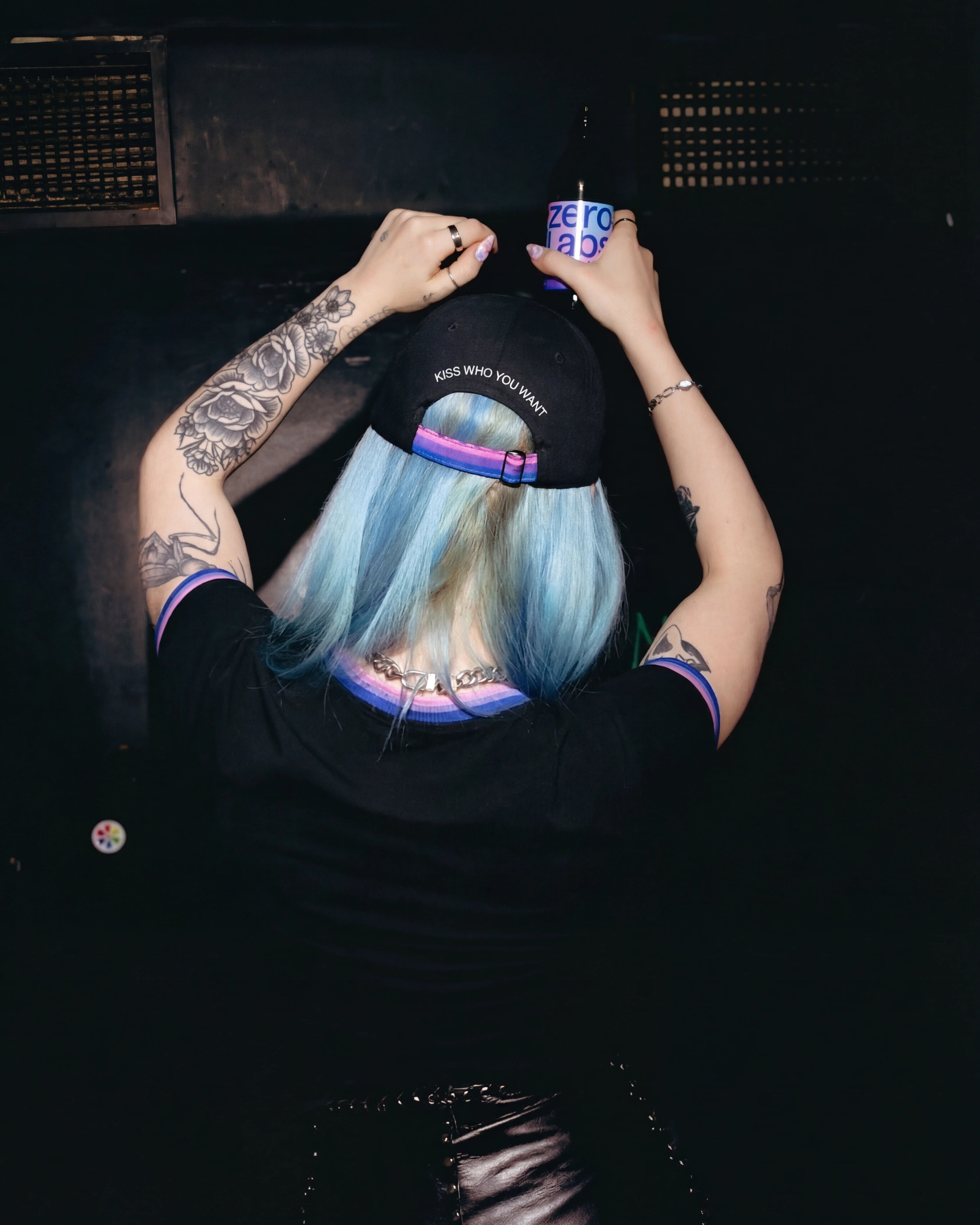 Model wearing a black Biweekly cap with bi pride flag strap and ‘Kiss Who You Want’ text on the back, back view, in a club scenario, dancing