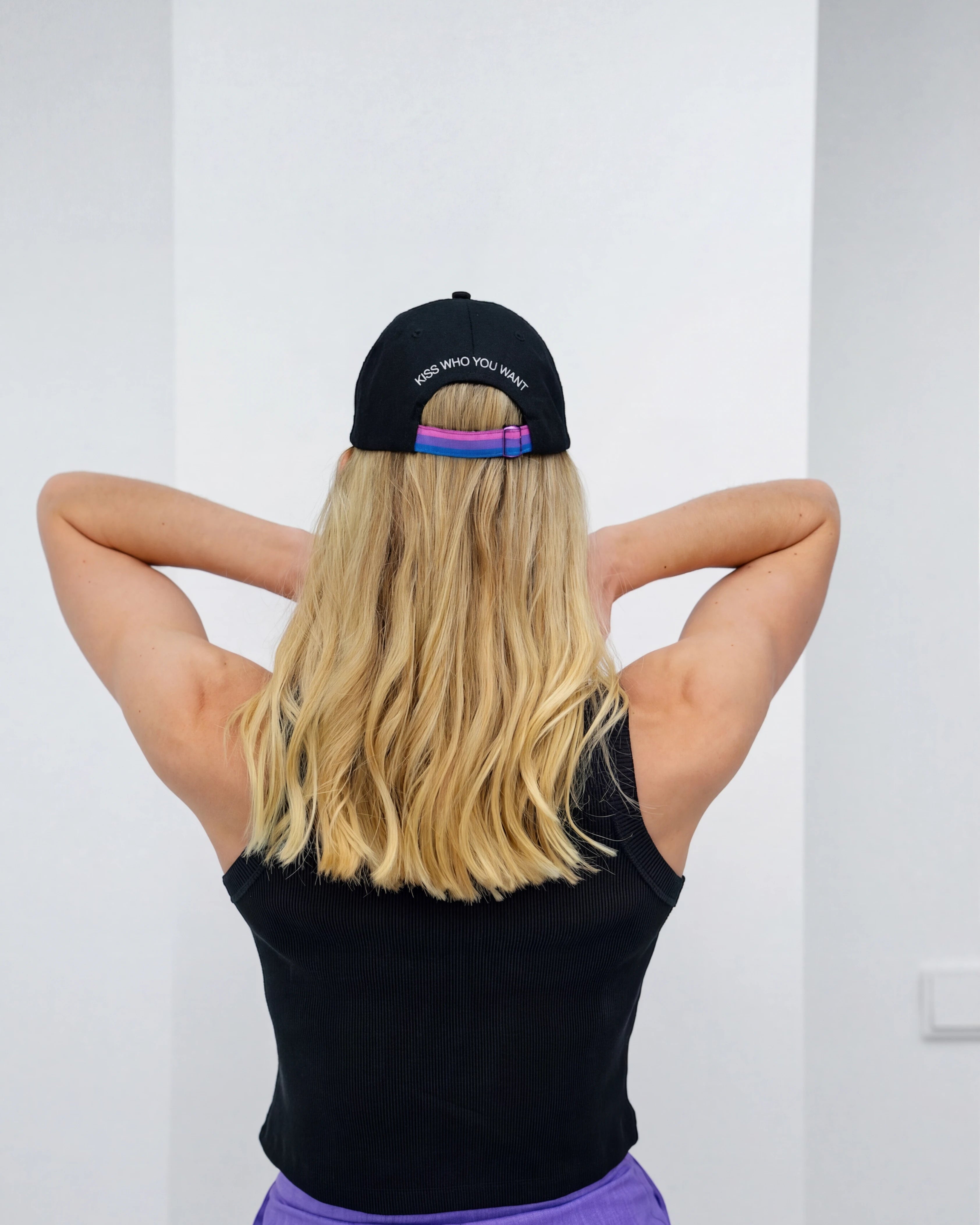 Model wearing a black Biweekly cap with bi pride flag strap and ‘Kiss Who You Want’ text on the back, back view against white background