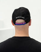 Detailshot of model wearing a black Biweekly cap with a strap in the colors of the bi pride flag and the text ‘Kiss Who You Want’ on the back against a white background
