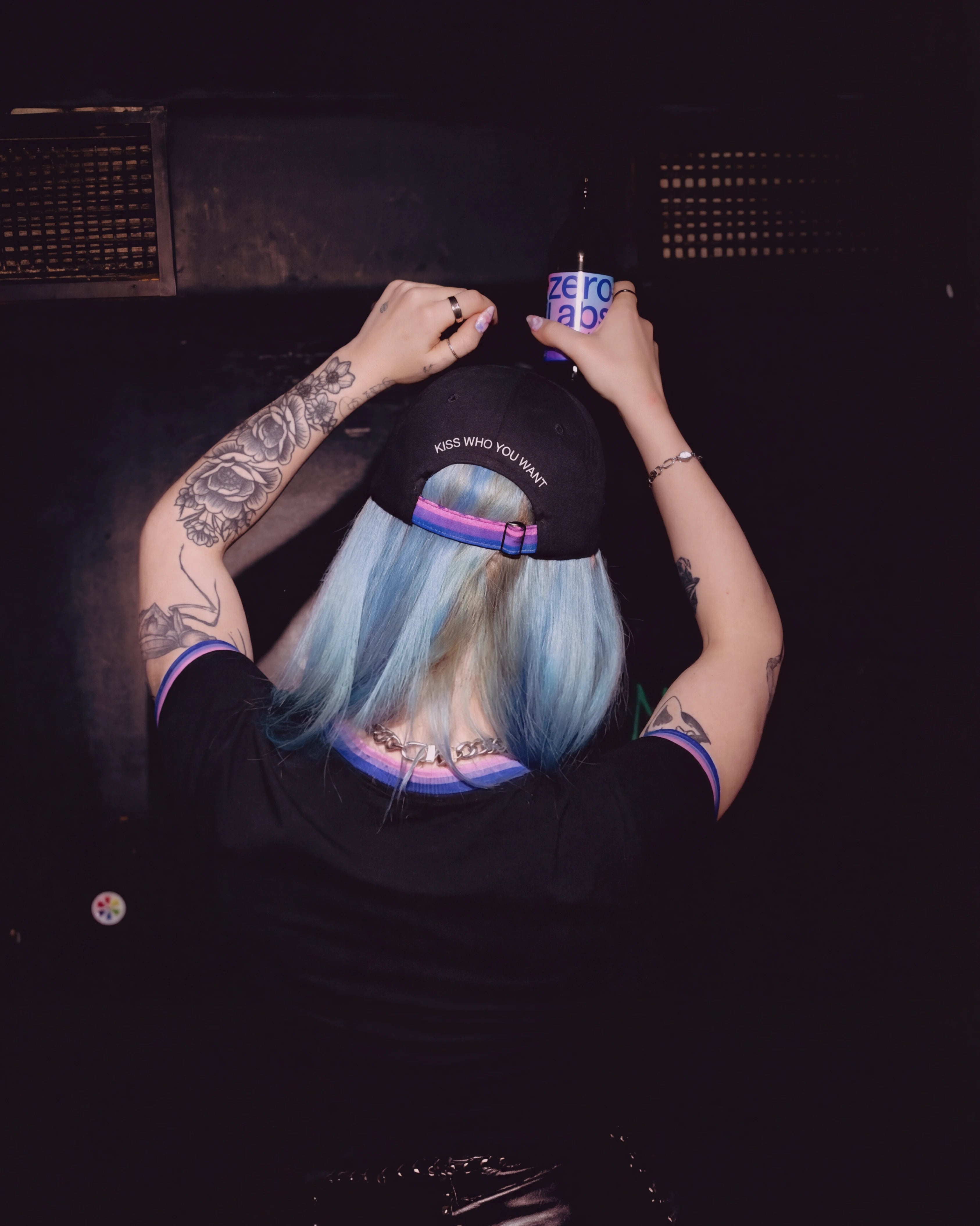 Model wearing a black Biweekly cap with bi pride flag strap and ‘Kiss Who You Want’ text on the back, back view, in a club scenario, dancing