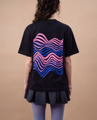 Model wears an oversized black Biweekly T-shirt with wavy bi pride flag graphic print and `Perfectly Not Straight` text on the print, back view against a beige background