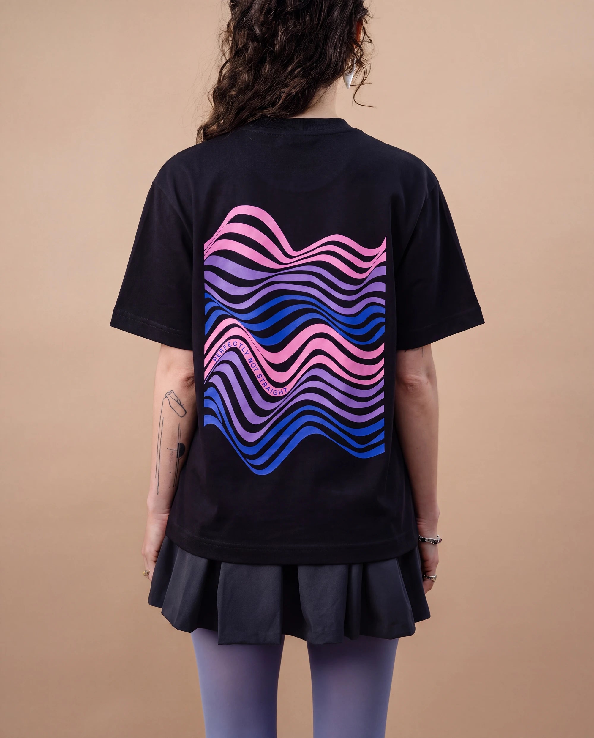 Model wears an oversized black Biweekly T-shirt with wavy bi pride flag graphic print and `Perfectly Not Straight` text on the print, back view against a beige background