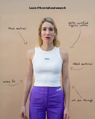 Model wearing a white Biweekly tank top with blue ‘Phase’ text that is crossed out, front view against a beige background. Text that gives information about top. 